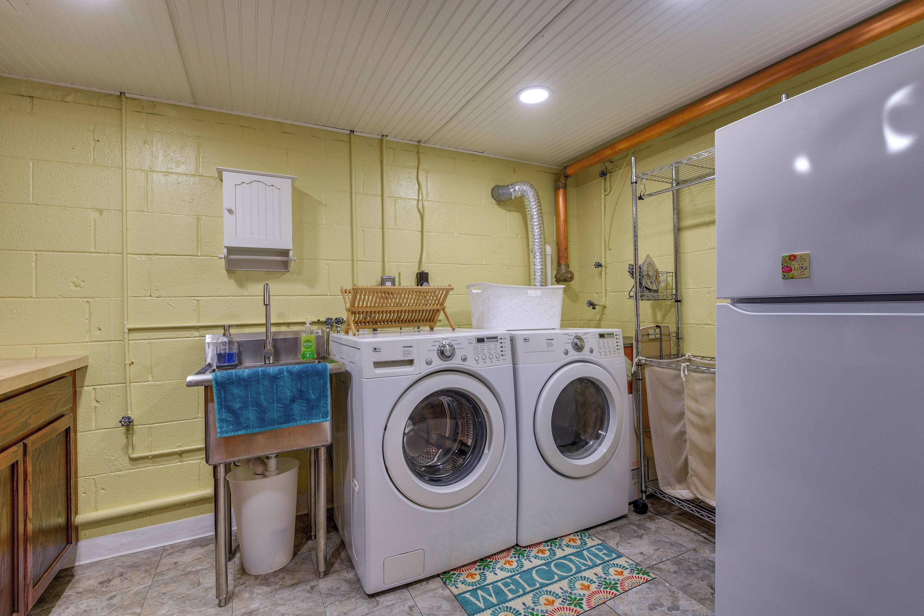 Une buanderie équipée de deux machines à laver à chargement frontal, d’un évier avec une serviette bleue, d’une armoire blanche et d’un paillasson.
