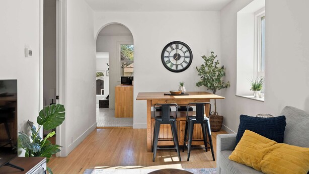 A modern living room with a wooden dining table, two black chairs, a grey sofa with yellow and blue cushions, and a large wall clock.