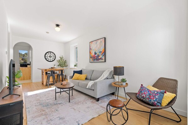 A modern living room with a grey sofa, a round coffee table, a dining area with a wooden table and chairs, and a large abstract painting on the wall. - The Sunny | 2 Suites | 10 Pax | Pet Friendly Stay! (Calgary)