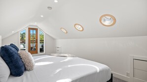 Una camera da letto con soffitto a falde, un letto con biancheria bianca, due applique tonde e vista su uno spazio esterno attraverso una porta.