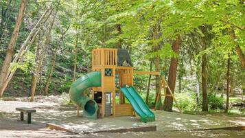 A wooden play structure with a slide and a climbing wall in a wooded area.