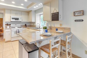Uma cozinha moderna com armários brancos, micro-ondas embutido, geladeira de aço inoxidável e bancada de granito. Há banquetas e um quadro negro com escrita na parede.