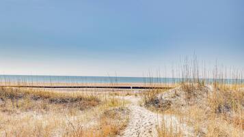 Un sentier de sable qui longe la plage et mène à l’océan, avec une promenade en bois visible au loin.