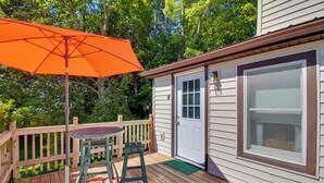 Une terrasse en bois avec une table et un parasol orange, une porte blanche et une fenêtre avec des stores.