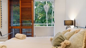 A bedroom with a bed, a window with wooden shutters, a lamp, and a view of tropical foliage.