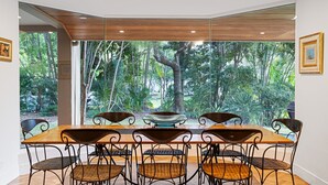 A dining area with a wooden table and wicker chairs, large windows with a view of a forest.