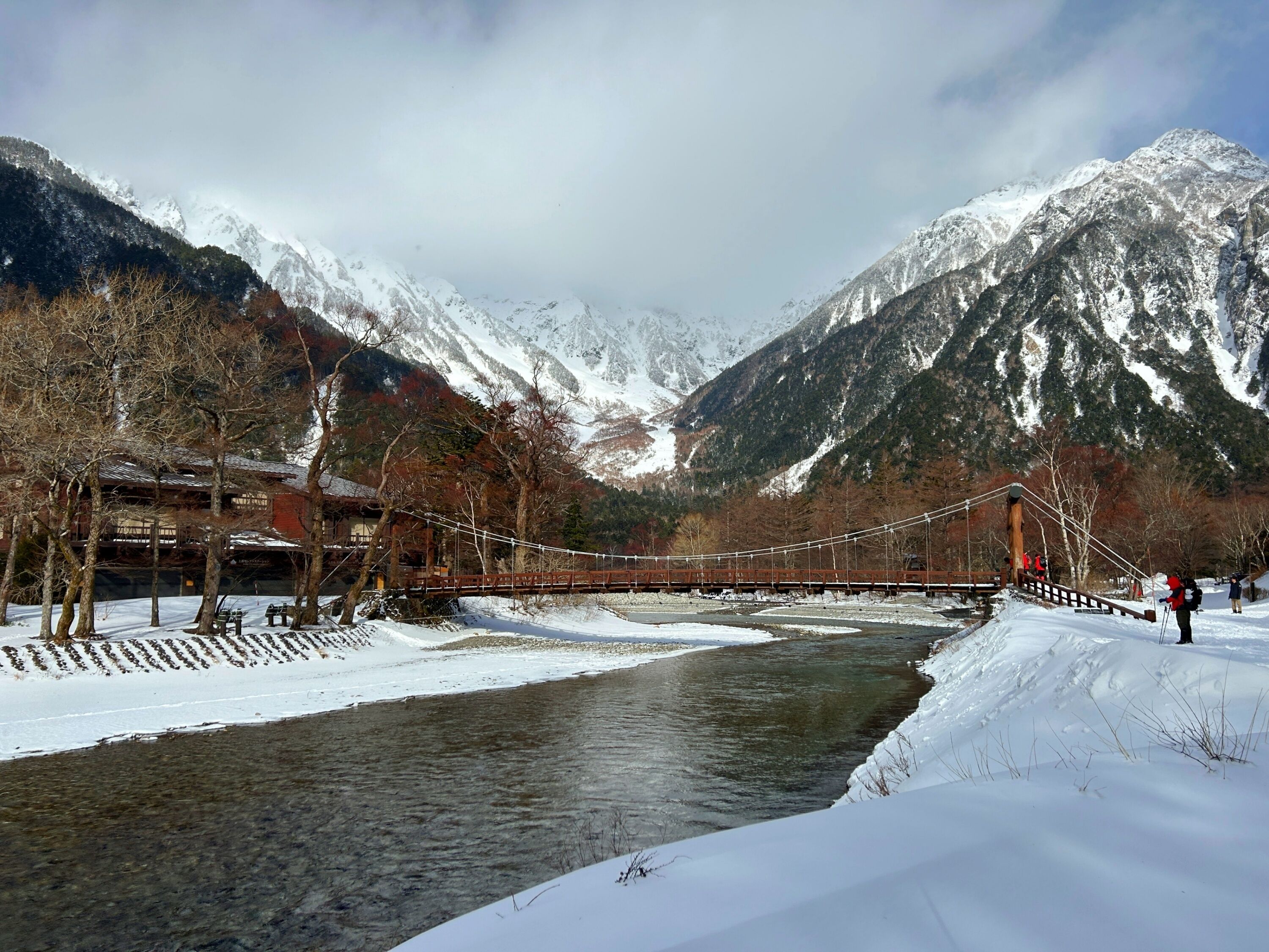 A snow-covered landscape with a wooden bridge over a river, mountains in the background, and a building with a red roof.