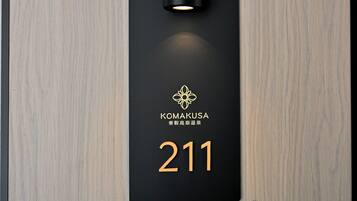 Ein schwarzes Schild mit der Nummer 211 und dem Namen "KOMAKUSA" in goldener Schrift, an einer Holzwand befestigt.