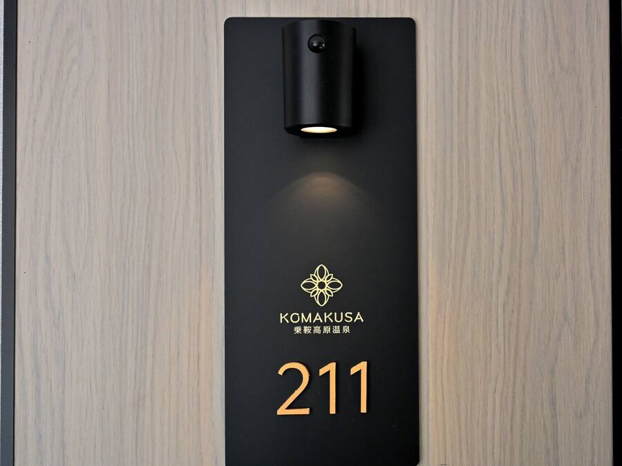 Ein schwarzes Schild mit der Nummer 211 und dem Namen "KOMAKUSA" in goldener Schrift, an einer Holzwand befestigt.