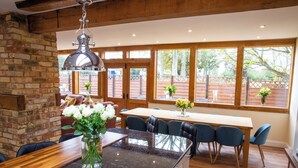 A kitchen with a wooden island, a dining table, and chairs. There is a brick wall and large windows with a view of trees. - Chapel Barn (Bedford)