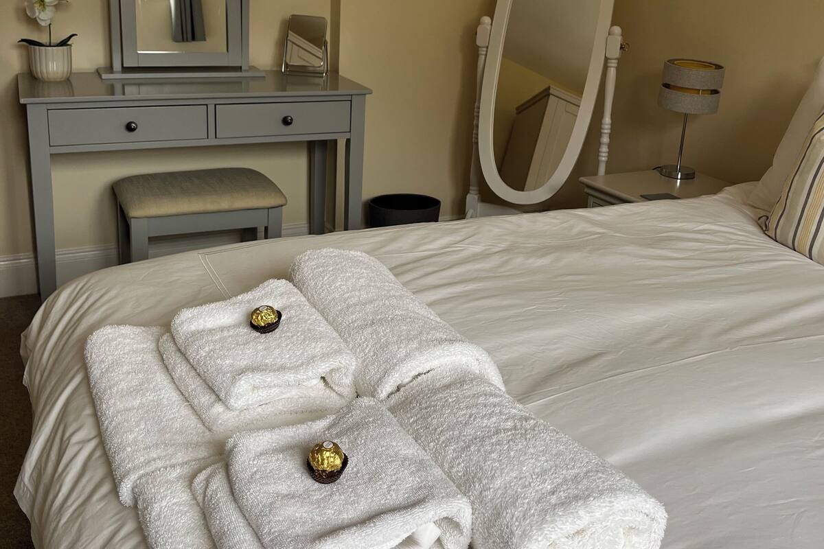 Una habitación de hotel con cama, tocador con espejo, banqueta y lámpara.