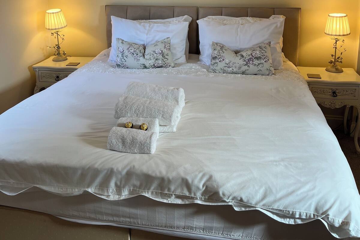 Una cama bien hecha con sábanas blancas y almohadas con motivos florales, una araña de cristal sobre ella y un cuadro en la pared.