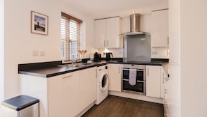 A compact kitchen with white cabinets, a stainless steel range hood, a built-in oven, a washing machine, and a window with blinds.