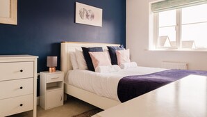 A bedroom with a bed, bedside tables, a nightstand, a window with blinds, and a wall-mounted picture. - Beautiful 3 Bedroom Home Near Worcester (Kempsey)
