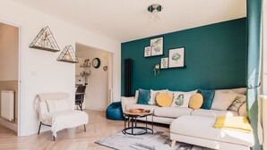 A modern living room with a teal accent wall, a white sofa with yellow and blue cushions, a white armchair, a coffee table, and a dining area visible in the background. - Beautiful 3 Bedroom Home Near Worcester (Kempsey)