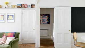 A cozy living room with a green sofa, a small wooden table, and a wicker chair. There are white doors leading to another room, and a colorful artwork on the wall. - Beautiful 3 Bedroom Home in Bristol (Bristol)