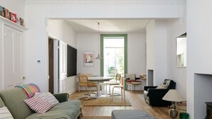 A living room with a green sofa, a grey ottoman, a dining table with chairs, a black armchair, and a fireplace. - Beautiful 3 Bedroom Home in Bristol (Bristol)