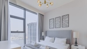 A modern hotel room with a large bed, a desk, and a view of a cityscape through large windows. - Creek Rise Residences - Creek Harbour (Dubai)