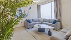 A modern living room with a grey sectional sofa, a round coffee table, and a large window with curtains. - Creek Rise Residences - Creek Harbour (Dubai)