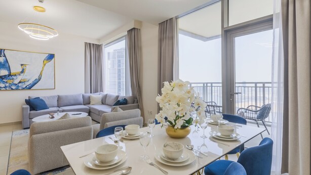 A modern living room with a dining area, grey sofas, a white dining table set, and a balcony with a view. - Creek Rise Residences - Creek Harbour (Dubai)