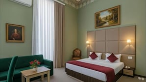 A hotel room with a bed, a green sofa, a small table with a vase of flowers, a painting on the wall, and a window with curtains. - Bella Garden Hotel (Baku)