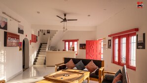 A spacious living area with a wooden coffee table, a sofa with cushions, a red-framed window, and a staircase leading to an upper floor. - Moustache Hostel Bir (Baijnath)