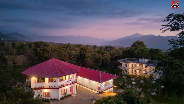 A two-story building with a red roof, surrounded by greenery and mountains in the background. - Moustache Hostel Bir (Baijnath)