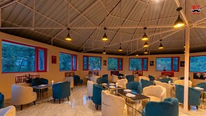 A spacious indoor area with multiple seating arrangements, large windows, and a high ceiling with exposed beams. - Moustache Hostel Bir (Baijnath)