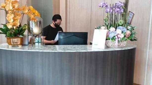 Ένας πάγκος υποδοχής με ένα άτομο καθισμένο πίσω του, έναν φορητό υπολογιστή και δύο διακοσμητικά σύνθετα με λουλούδια.