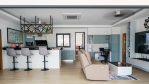 A modern living room with a sofa, a coffee table, and a TV. There is a kitchen area with a bar and stools. - Tilawan Villa - Cozy 3 Bedrooms Pool Villa in Chalong (Chang Wat Phuket)