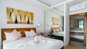 A hotel room with a large bed, a wooden headboard, a painting on the wall, a mirror, and a bathroom visible through an open door. - Tilawan Villa - Cozy 3 Bedrooms Pool Villa in Chalong (Chang Wat Phuket)