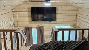 Ein Holzblockhaus-Interieur mit einem an der Wand montierten Flachbildfernseher, einem Deckenventilator und einem Balkon mit Blick ins Freie.