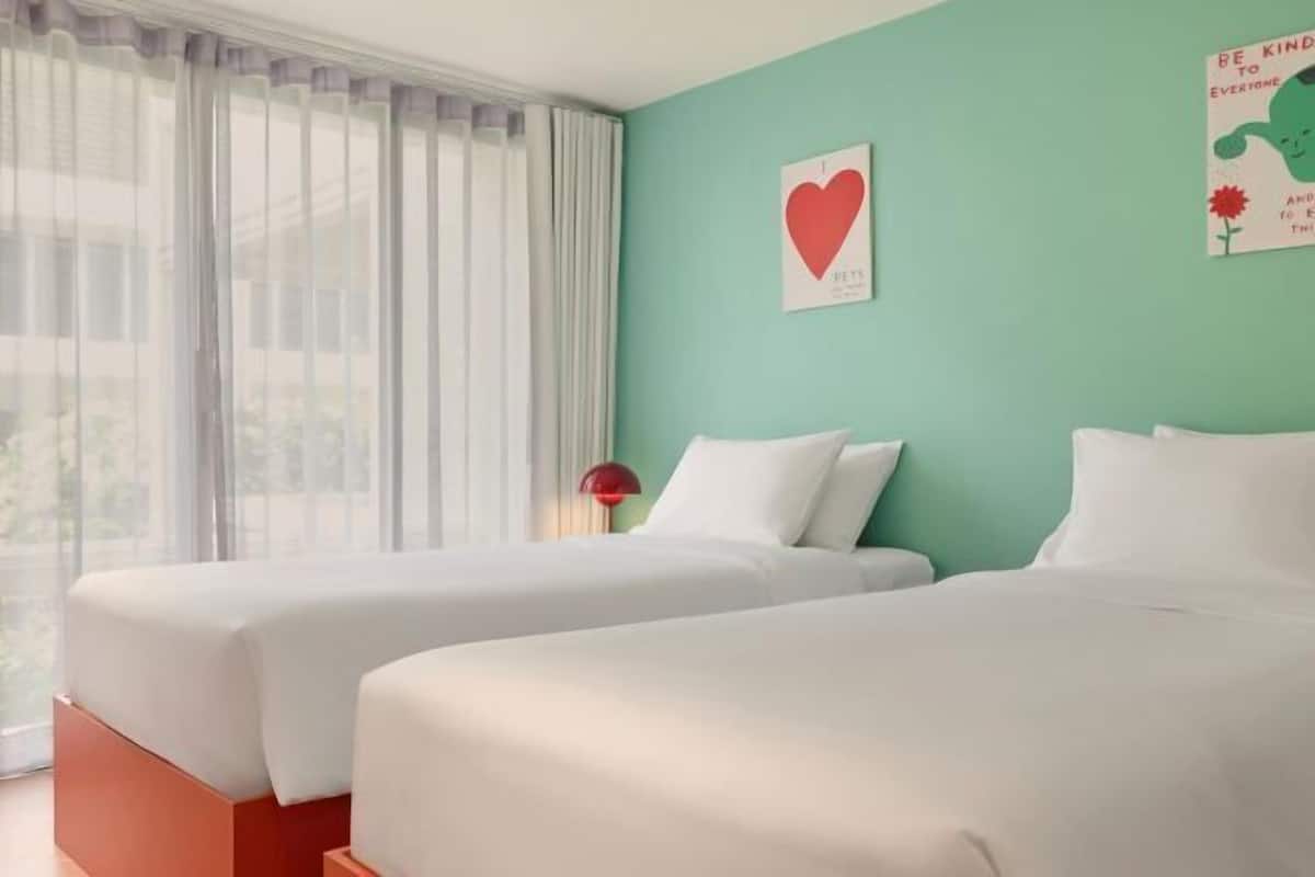 Une chambre d’hôtel avec deux lits, un mur de couleur turquoise et une fenêtre ornée de rideaux légers.