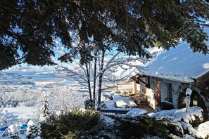 Una cabaña de madera en la montaña, cubierta de nieve, con un porche y vistas a un paisaje nevado.
