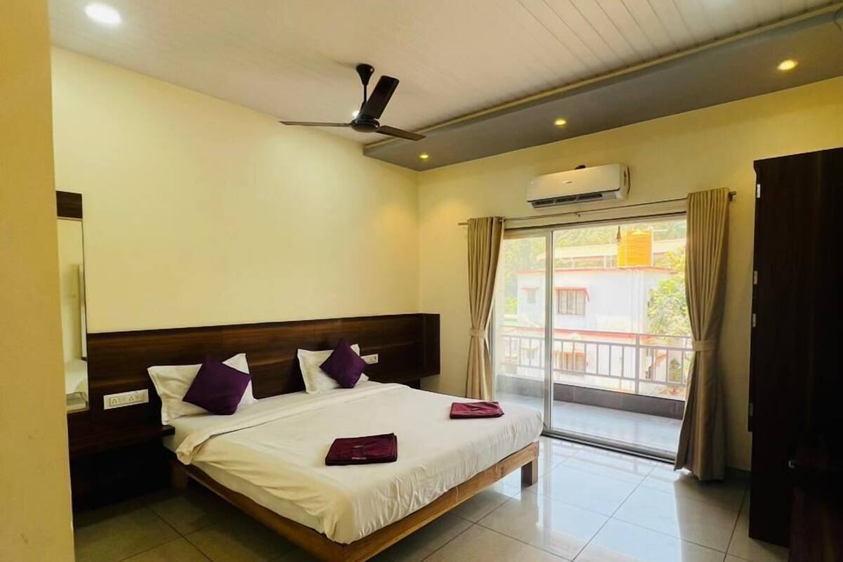 A hotel room with a bed, a ceiling fan, an air conditioning unit, and a view of the outside through a large window.