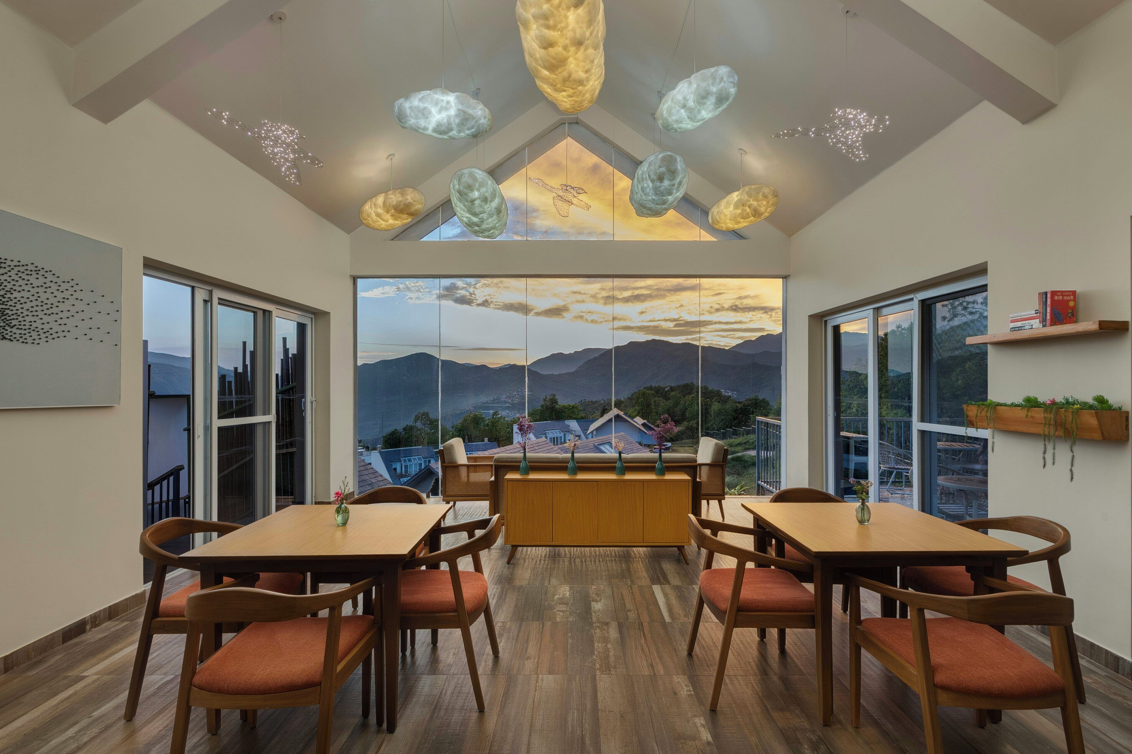 Un comedor con mesas y sillas de madera, un ventanal con vista a la montaña y lámparas colgantes.