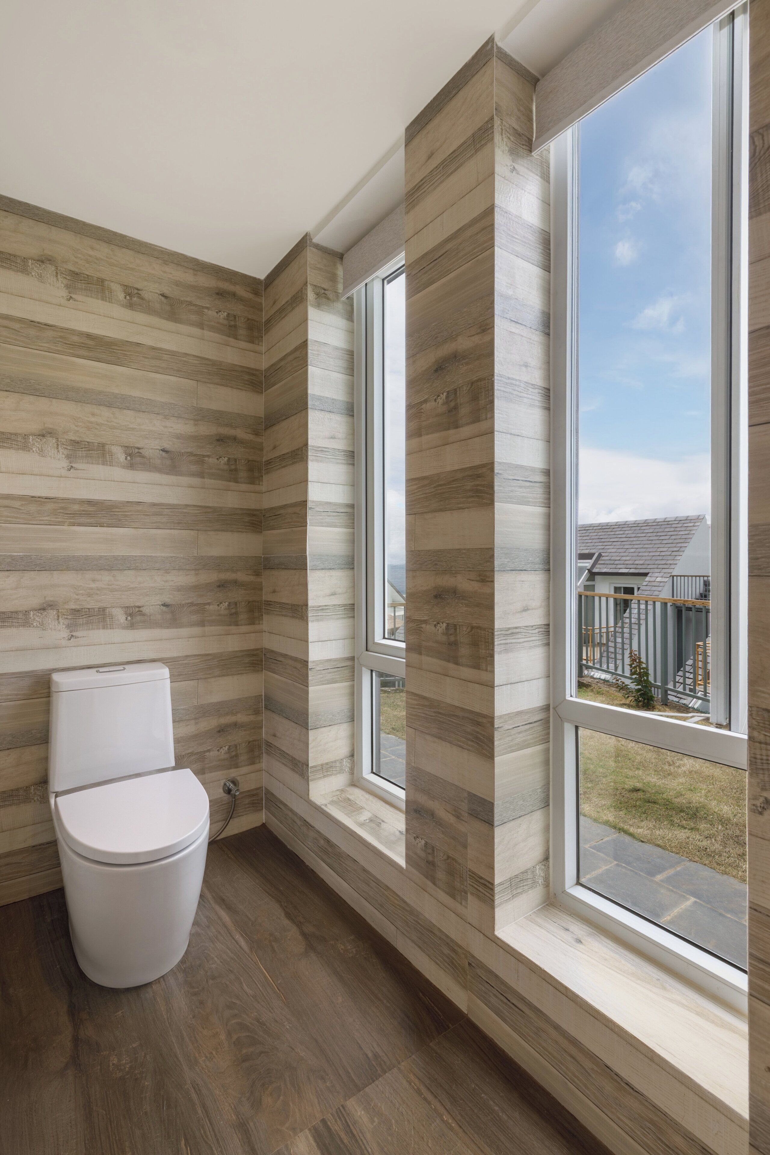 A bathroom with a toilet, wooden walls, and large windows offering a view of the outside.