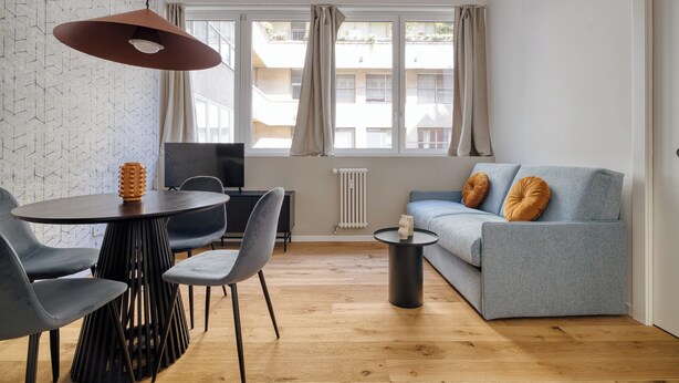 Egy modern nappali, melyben szürke kanapé, kék székek, egy kerek asztal és egy függesztett lámpa található.