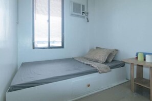 Een eenpersoonsbed met matras, kussens en dekbed. Er is een raam met zonwering, een klein tafeltje met een kopje en een wandairconditioner.