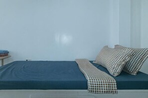 Una cama con colchón azul y almohadas a cuadros, una mesita con un objeto rojo y una pared blanca lisa.