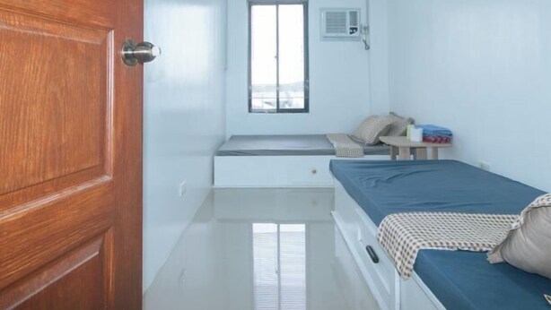 A small, clean room with a wooden door, a window with blinds, a bed with a blue mattress, and a small table with a chair. - Skybox Pension House by RedDoorz (Iloilo City)