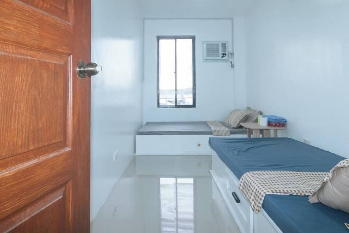 Une petite pièce propre, avec une porte en bois, une fenêtre avec des stores, un lit doté d’un matelas bleu et une petite table accompagnée d’une chaise.