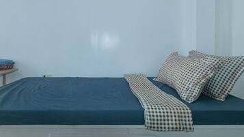 A bed with a blue mattress and patterned pillows, a bedside table with a book, and a plain wall.