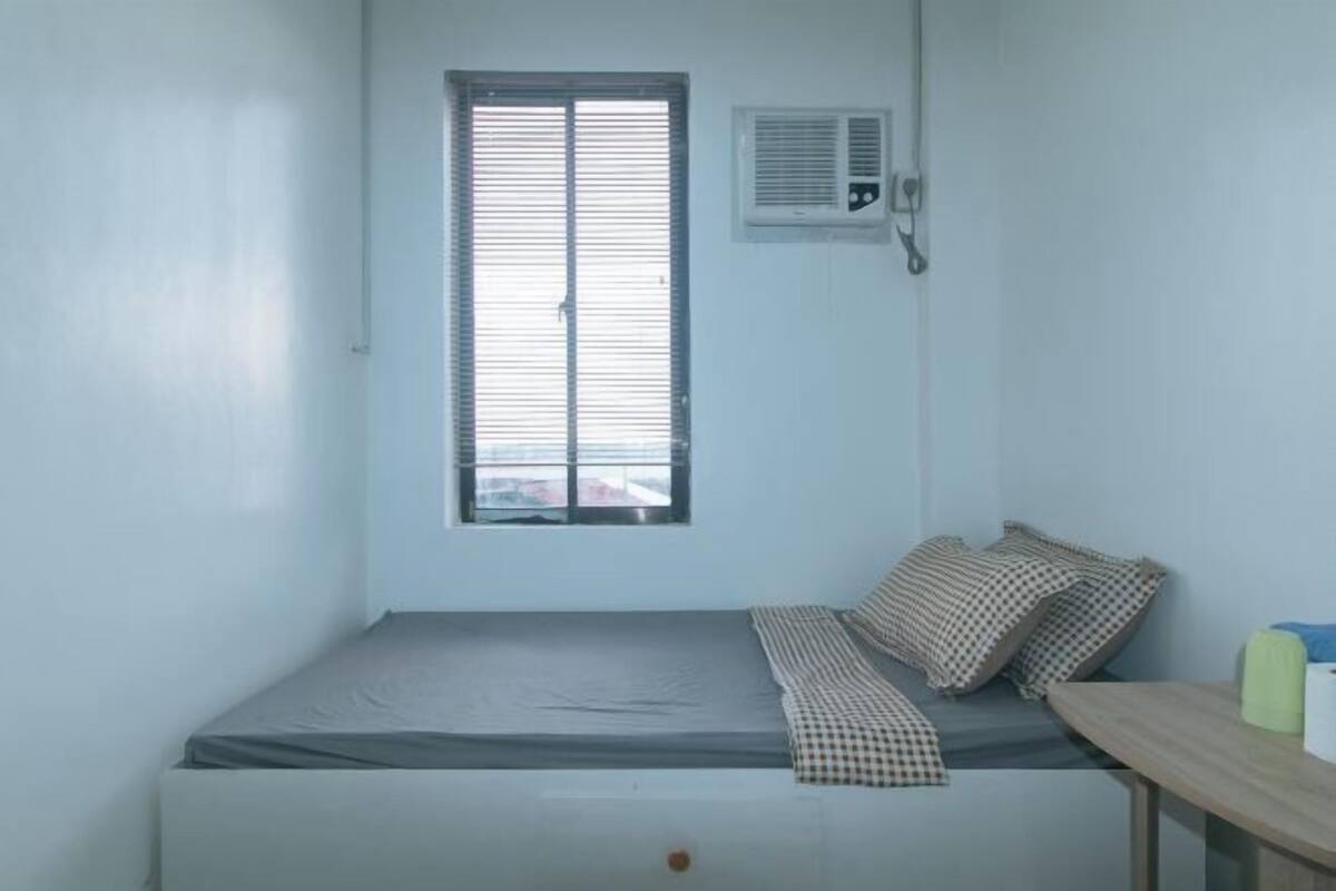 Un letto singolo con coperta a quadri, una finestra con persiane, un condizionatore d'aria e un tavolino con una bottiglia verde.