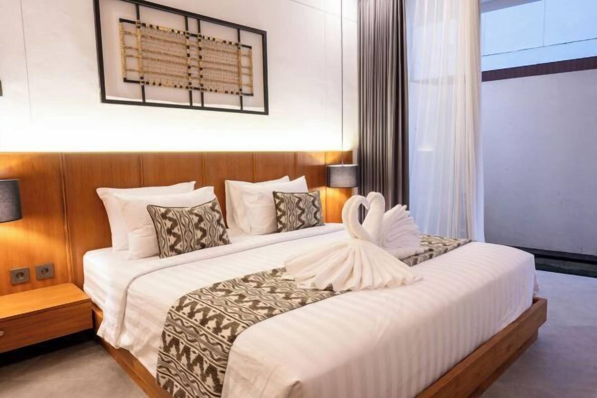 A modern hotel room with a wooden headboard, a large bed with white linens, a patterned throw blanket, and a wall-mounted artwork.