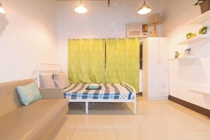 A room with a bed, a sofa, a radiator, a shelf with plants, and a window with curtains. - San Marino JY7 Rentals by RedDoorz (Cebu City)