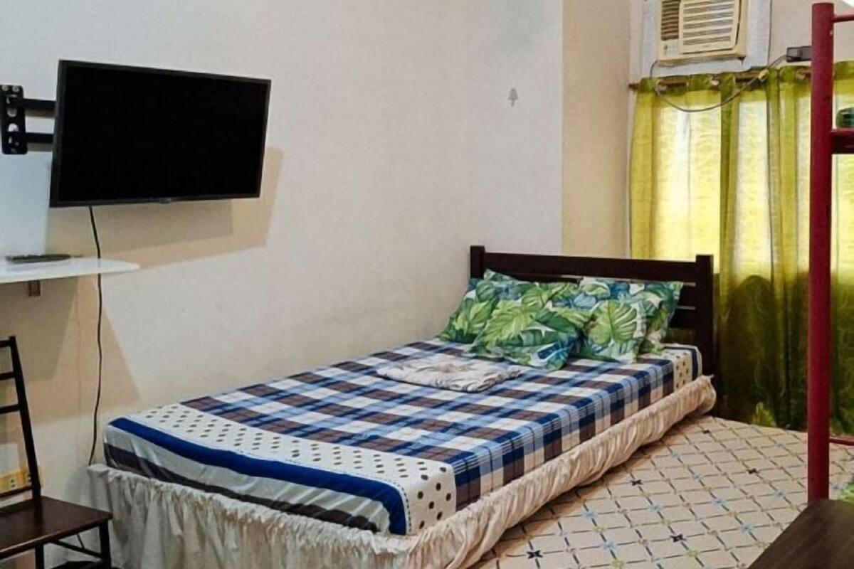Ein Einzelbett mit karierter Bettdecke, ein an der Wand befestigter Fernseher, ein Sessel und ein Fenster mit Vorhängen.