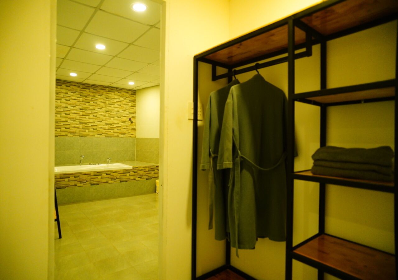 A bathroom with a tiled floor, a sink, and a shower area. There is a wooden shelf with folded towels and a robe hanging on it.