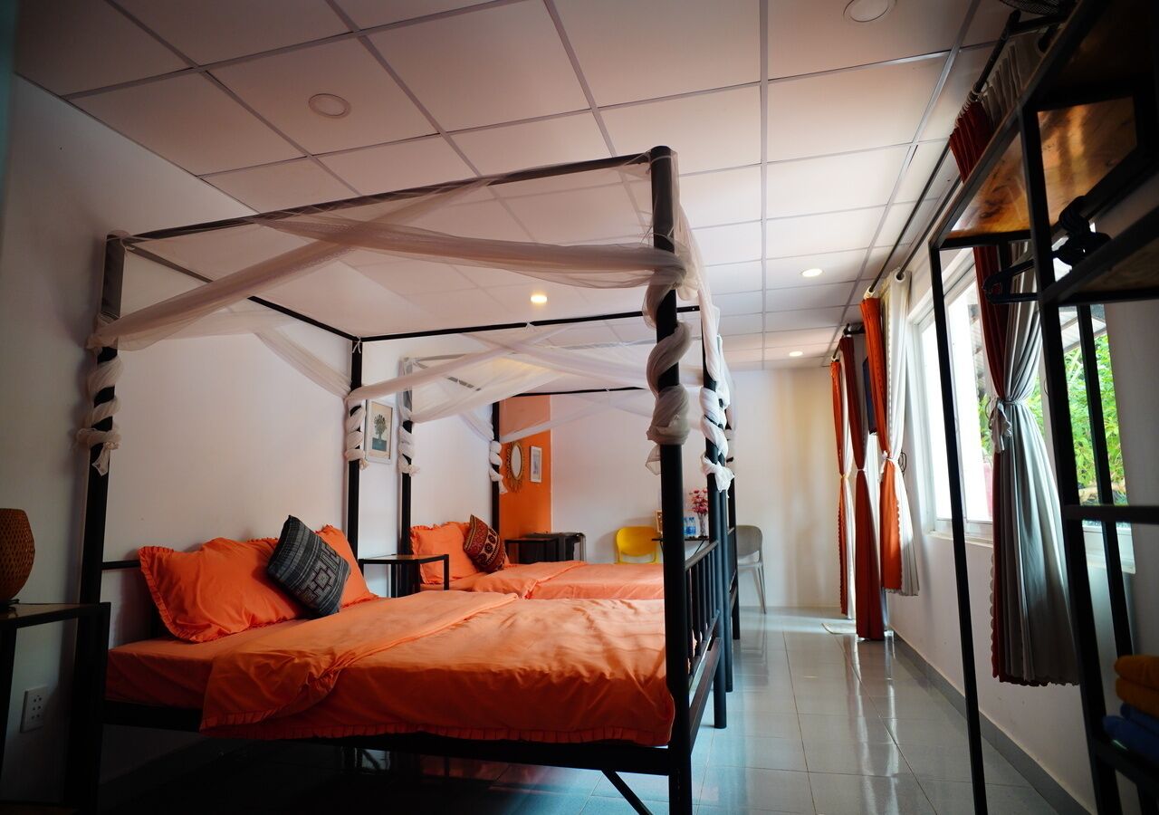 Ein Bett mit Baldachin, mit orangefarbener Bettwäsche, ein kleiner Tisch mit einem Stuhl und ein Fenster mit Vorhängen.