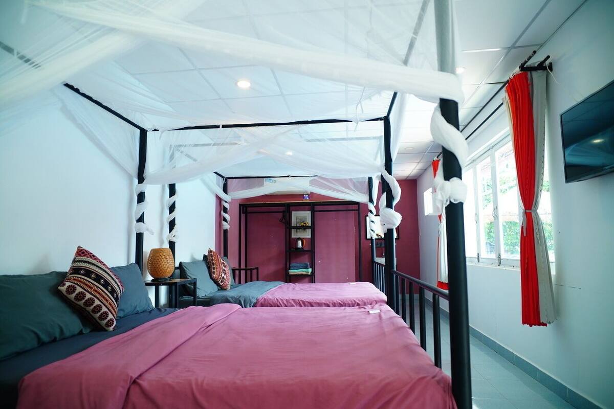Un dormitorio con cama, almohadas, una mesita de noche, una pared roja y una ventana con cortinas.